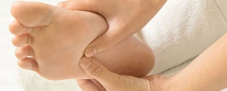 A person is massaging the sole of their bare foot with both hands seated on a white surface