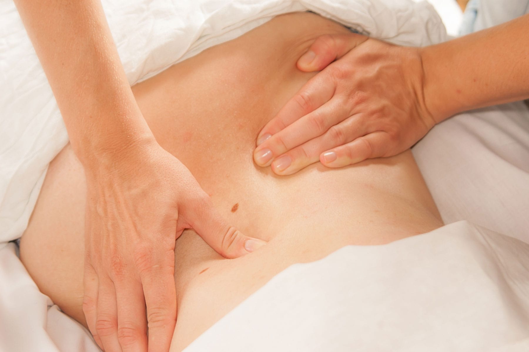 Close up of a persons back being massaged by a therapists hands with white sheets partially covering the person