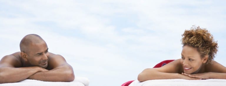 A man and a woman lie on sun loungers facing each other smiling and relaxing under a clear sky