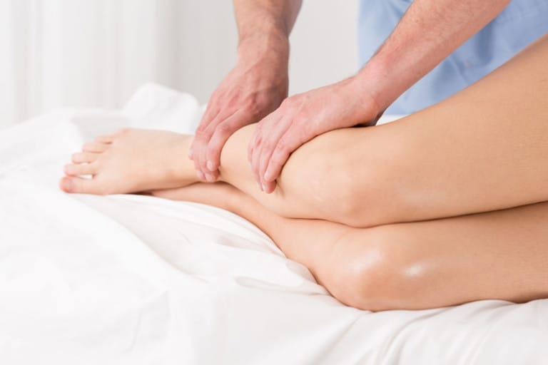 A person is lying on a bed receiving a leg massage Another persons hands are massaging the lower legs and ankles Both individuals faces are not visible in the image
