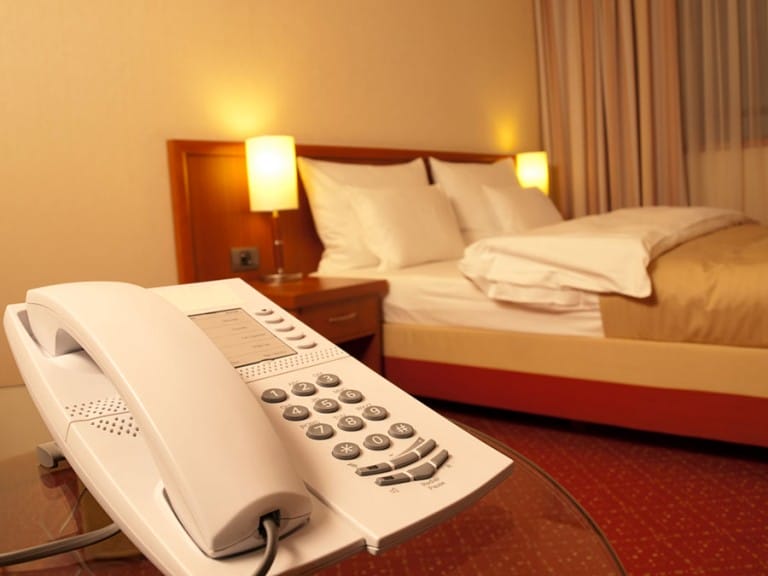 A hotel room with a neatly made bed side lamps on nightstands and a white telephone on a glass topped desk in the foreground