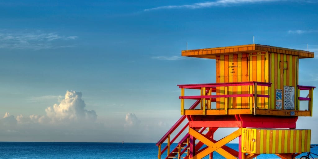 A brightly colored yellow and red lifeguard tower stands near the shore with a clear blue sky and calm ocean in the background