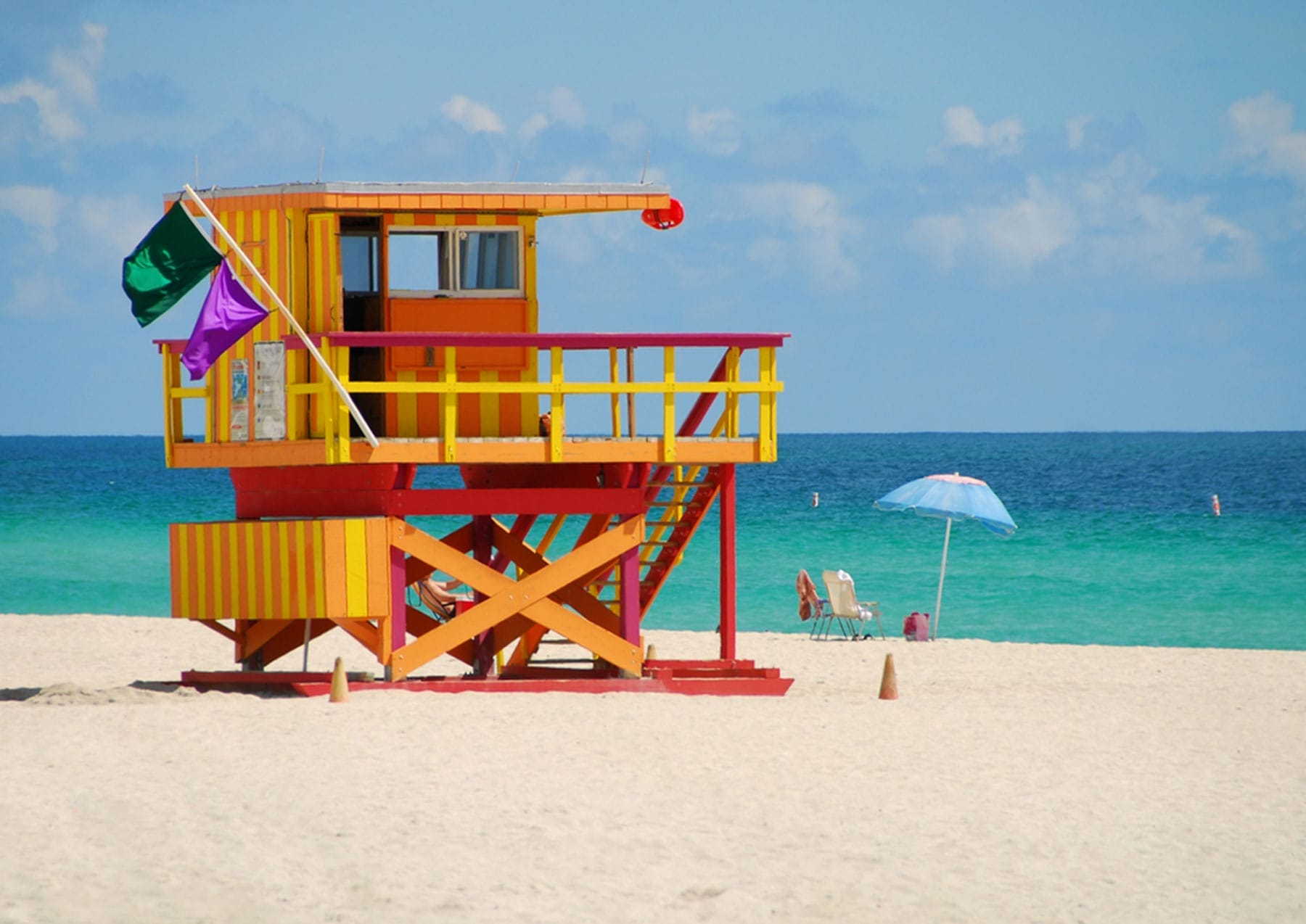 A colorful lifeguard tower stands on a sandy beach with calm turquoise water and a partly cloudy sky Nearby an umbrella and two beach chairs sit close to the water