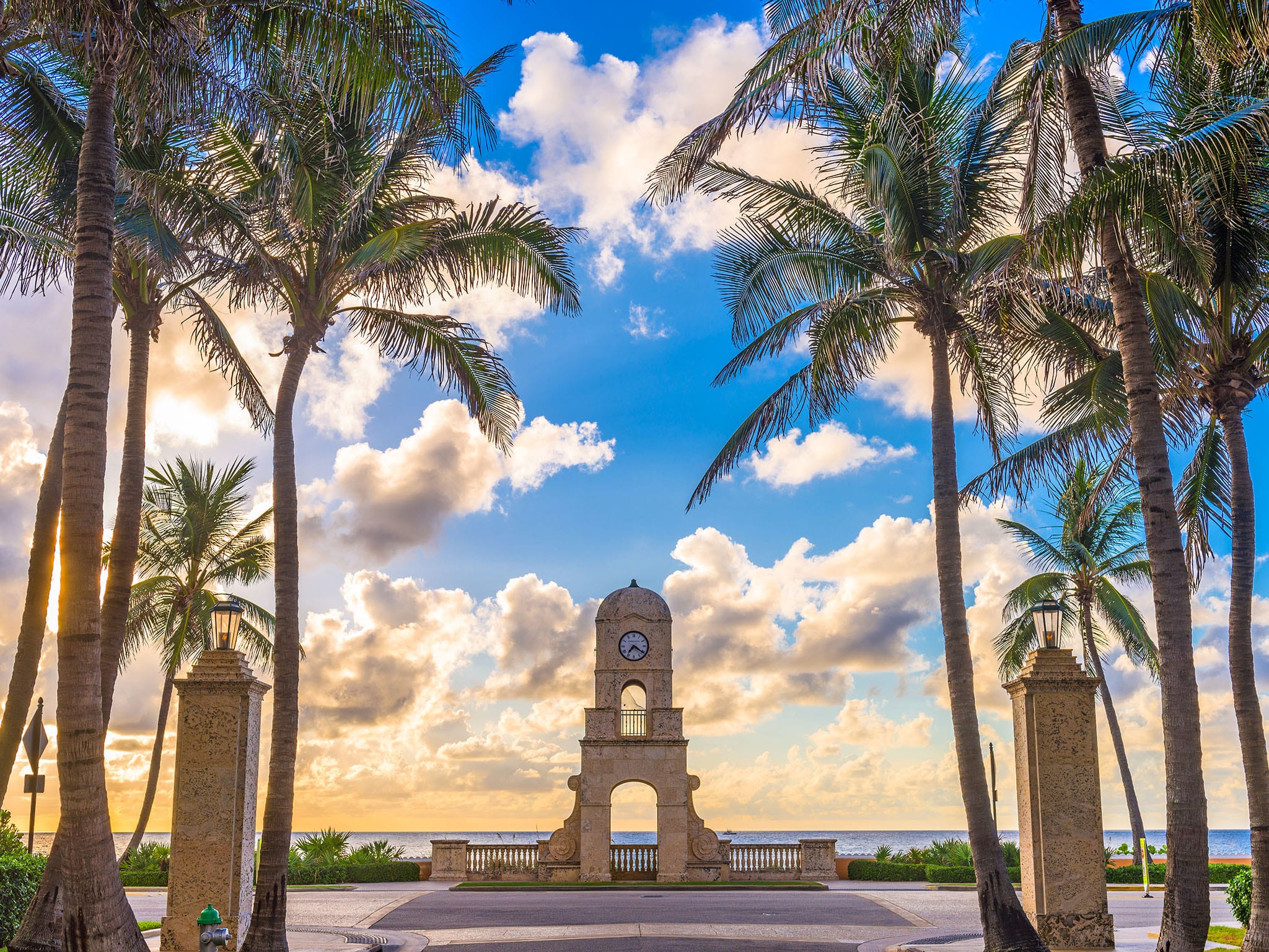 A clock tower framed by palm trees stands against a backdrop of a vibrant partly cloudy sky and the ocean at sunset