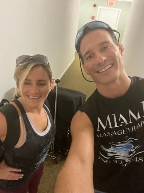 Two people are standing in a hallway both smiling The person on the left is wearing sunglasses on their head and a tank top with a backpack The person on the right is wearing a sleeveless shirt and has a travel cart