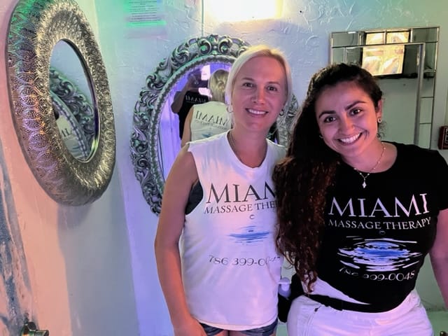 Two women in Miami Massage Therapy shirts stand in front of ornate mirrors smiling at the camera One wears a white shirt and the other a black shirt