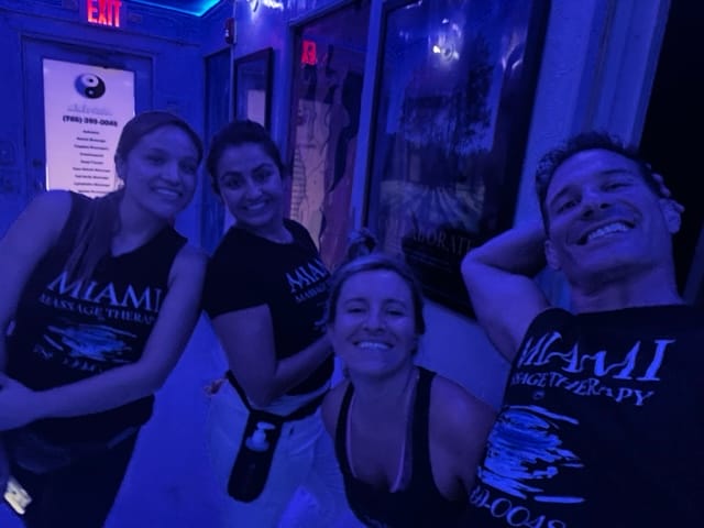 Four smiling people wearing MIAMI Massage Therapy shirts pose together in a dimly lit blue tinted room with posters on the walls