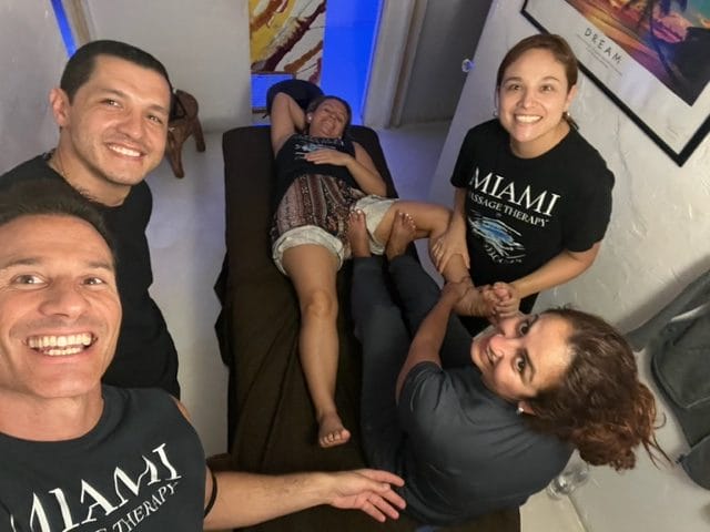 Four people smile at the camera while a woman lies on a massage table receiving a foot massage Two of the people wear Miami Massage Therapy T shirts
