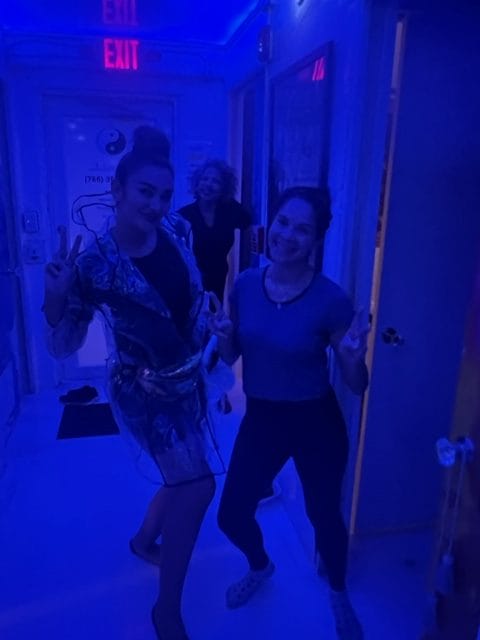 Three people pose in a hallway lit by blue lighting with two in the front giving peace signs and a person in the back smiling