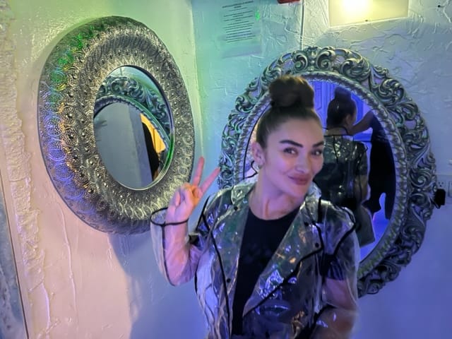 A person wearing a shiny jacket poses with a peace sign in front of a set of decorative mirrors on a textured wall