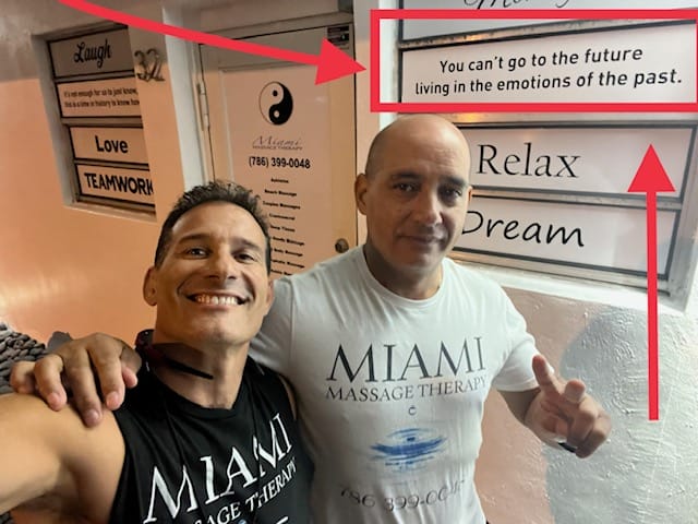 Two men in MIAMI Massage Therapy shirts pose in front of a wall with motivational signs including one that reads You cant go to the future living in the emotions of the past
