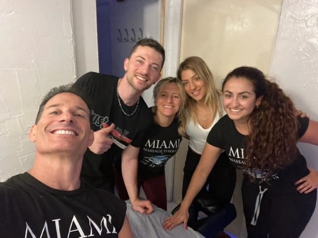 Five people in black shirts pose and smile in a room Four are standing and one is taking the photo The shirts read MIAMI and MASSAGE THERAPY