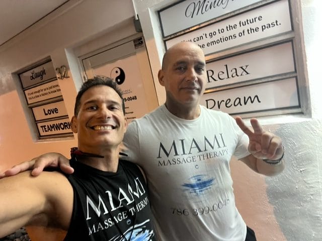 Two men standing side by side smiling both wearing Miami Massage Therapy T shirts One man is showing a peace sign and behind them is a wall with motivational text