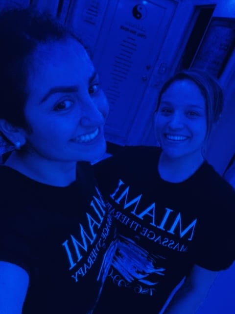 Two smiling women wearing black MIAMI t shirts pose for a selfie in a dimly lit blue tinted room A door and posters are visible in the background