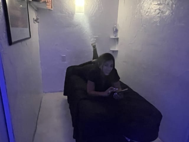 A person lies on a black sheet covered bed in a small dimly lit room holding a phone Walls are white and textured A water bottle is on a small shelf A framed picture hangs on the left wall