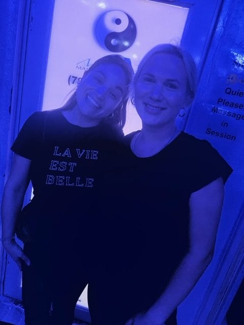 Two people stand smiling in front of a door with a Yin Yang symbol The person on the left wears a shirt that says LA VIE EST BELLE The scene is illuminated with blue lighting