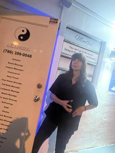 A woman stands outside a massage therapy center at night next to a sign with a quote and a door listing massage services