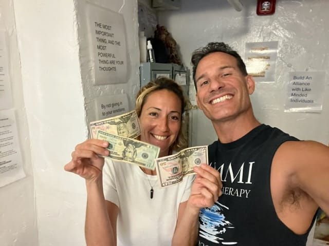 Two people smiling and holding various denominations of US currency in a room with motivational posters on the walls