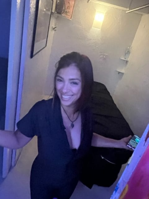 A woman with dark hair dressed in a black outfit smiles while standing in a small dimly lit room with white walls she holds a phone in her left hand