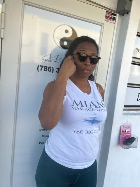 A woman wearing sunglasses and a Miami Massage Therapy tank top stands in front of a door with the same logo and a yin yang symbol She is touching the side of her head with one hand