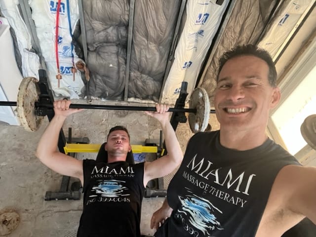 Two men wearing Miami Massage Therapy shirts are in an unfinished room One man is lifting weights on a bench press while the other takes a smiling selfie Building insulation is visible in the background