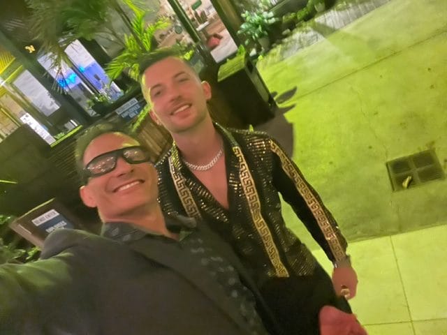 Two men posing for a selfie outdoors at night one wears a dark suit and glasses the other wears a stylish jacket with gold accents and a chain necklace