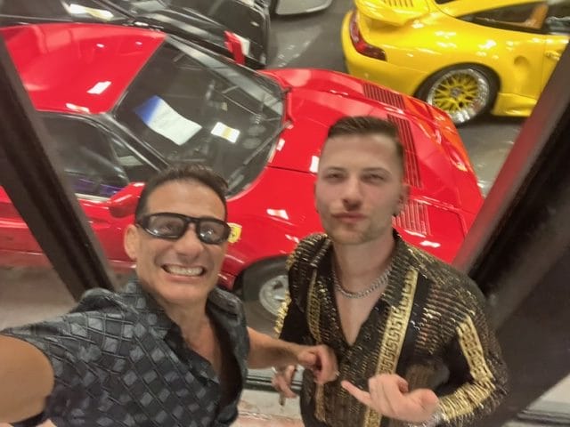 Two men take a selfie in front of high end sports cars one red and one yellow in an indoor display area