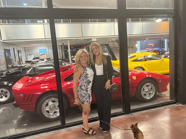 Two women stand smiling in front of a showroom window displaying two Ferrari cars one red and one yellow A small dog is visible in the foreground The setting appears to be indoors at night