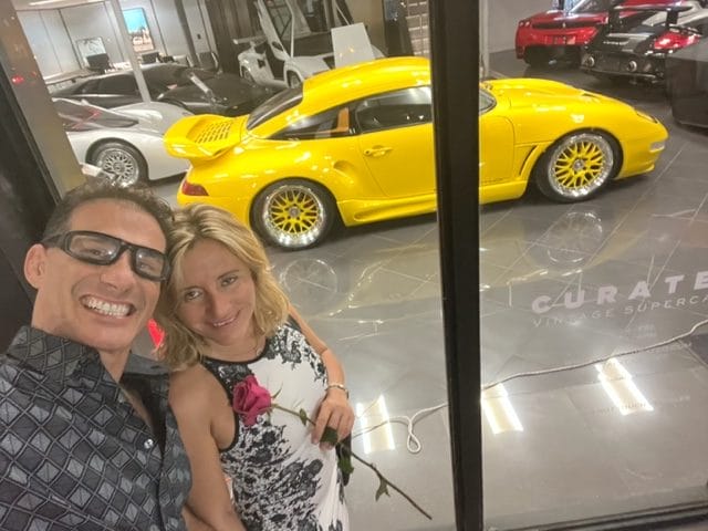 A smiling man and woman holding a rose pose in front of a display window showcasing various luxury cars including a prominent yellow car in the center