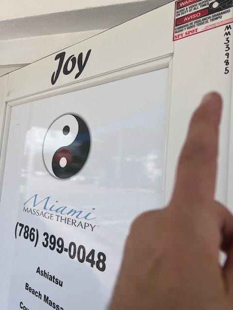 A finger points to a sign on a white door labeled Joy above a poster for Miami Massage Therapy The poster includes a yin yang symbol contact number and services like Ashiatsu and Beach Massage Therapy