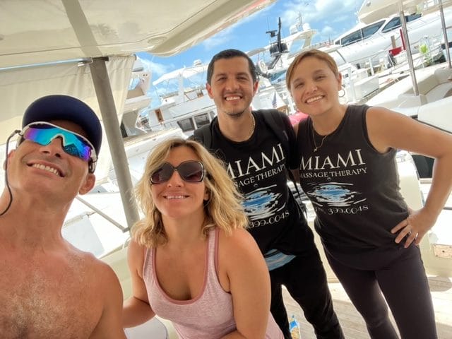 Four people are posing and smiling on a boat dock with yachts in the background Two of them are wearing black shirts reading Miami Massage Therapy It is a sunny day
