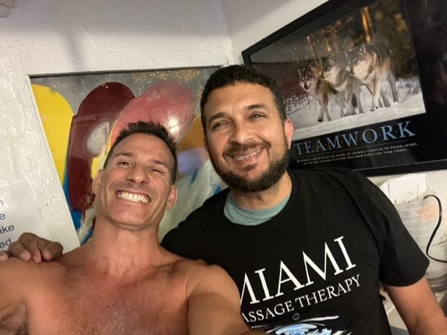 Two men are standing close together smiling at the camera One is shirtless and the other is wearing a Miami Massage Therapy shirt A poster with wolves and the word Teamwork is in the background