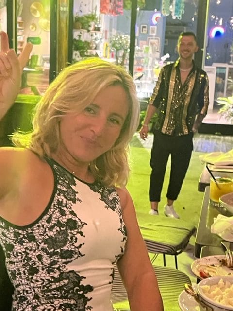A woman in a black and white dress smiles and makes a peace sign gesture at a restaurant with a man in the background wearing a sequined jacket and black pants