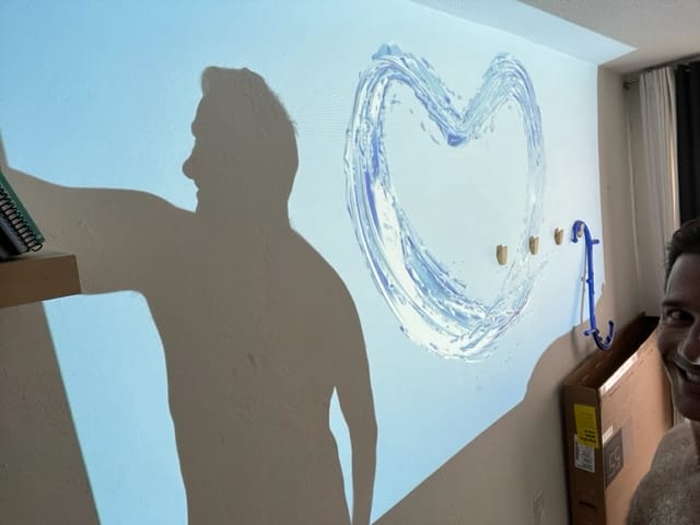 A mans shadow is projected on a wall while he stands next to a heart design Only part of his face is visible He appears to be attaching or adjusting something on the wall