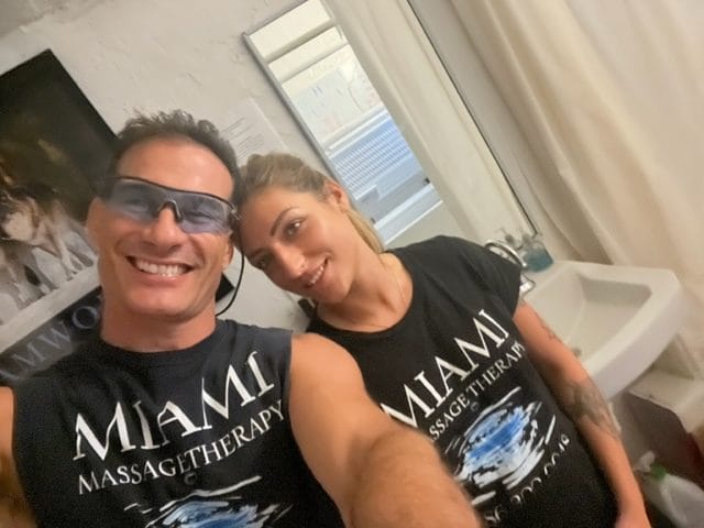 Two individuals smiling and posing together indoors both wearing black MIAMI Massage Therapy shirts One person is wearing protective glasses A sink and curtain are visible in the background