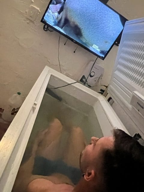 A person is lying in an ice bath inside a large cooler while watching a television mounted on the wall