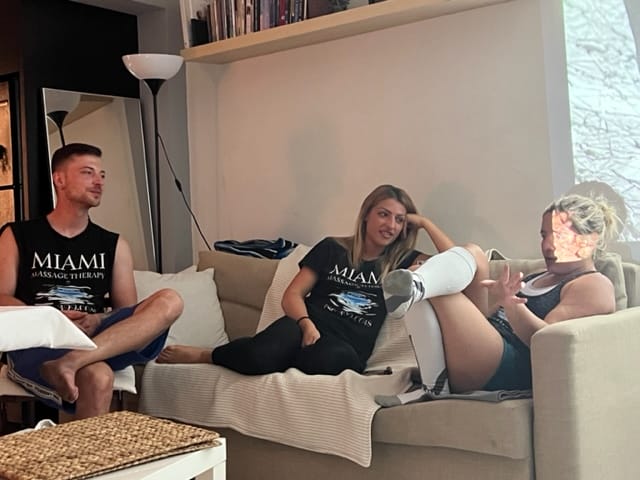 Three people are sitting on a couch in a living room engaged in conversation The person on the right has a leg elevated wearing a cast or bandage All are dressed casually in t shirts and shorts