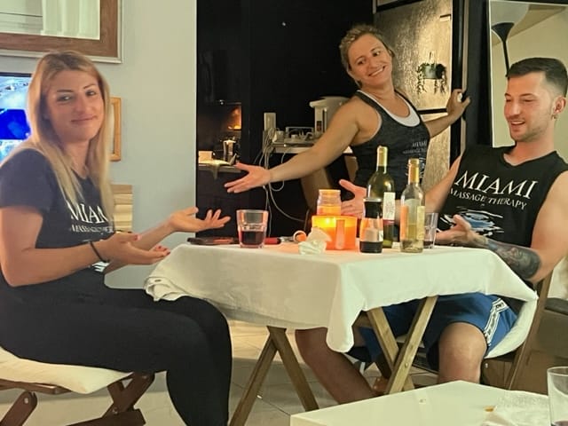 Three people in matching Miami Massage Therapy shirts are gathered around a table with drinks and snacks two sitting and one standing and gesturing with both arms