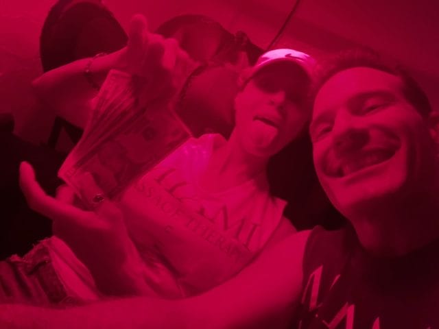 Two people take a selfie in red lighting One holds a fan of cash making a playful face with their tongue out while the other smiles at the camera