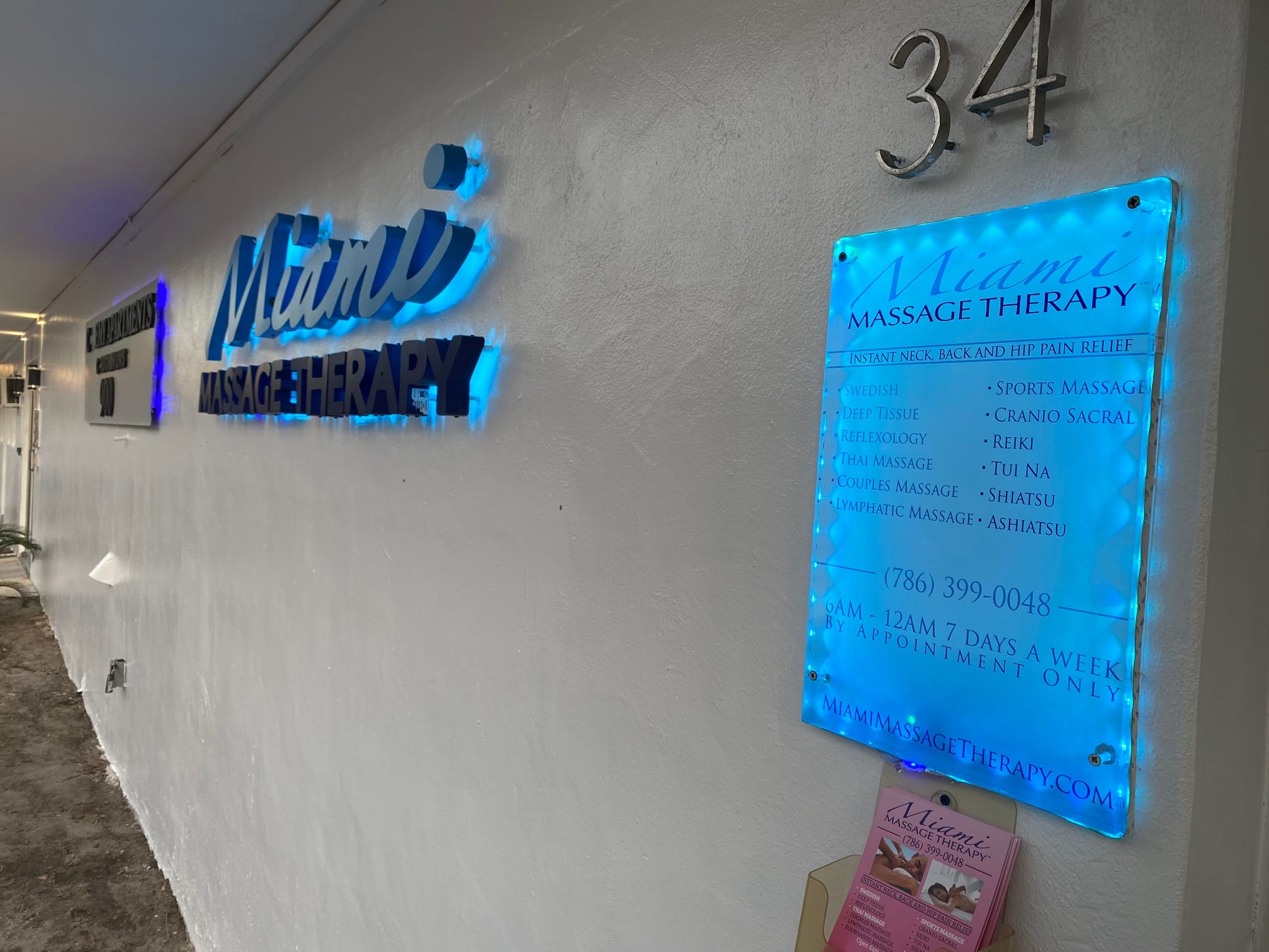 A white wall with a Miami Massage Therapy sign contact details and a list of services offered A business card holder is also visible The address number 34 is displayed above the signs