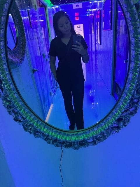A person wearing dark clothing takes a mirror selfie in a brightly lit blue hallway