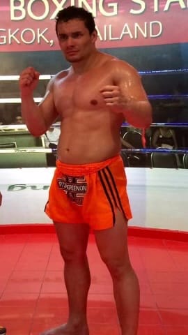 A man in orange shorts is standing in a boxing ring with his fists up posing for the camera The background displays text and an image of a boxing stadium in Thailand