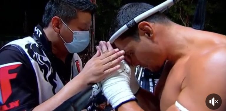 A fighter with a headband and wrapped hands bows his head pressing his palms together against those of a masked coach or trainer in a pre fight ritual