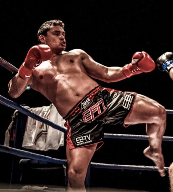 A male kickboxer in red gloves and black shorts delivering a high kick in a boxing ring during a match