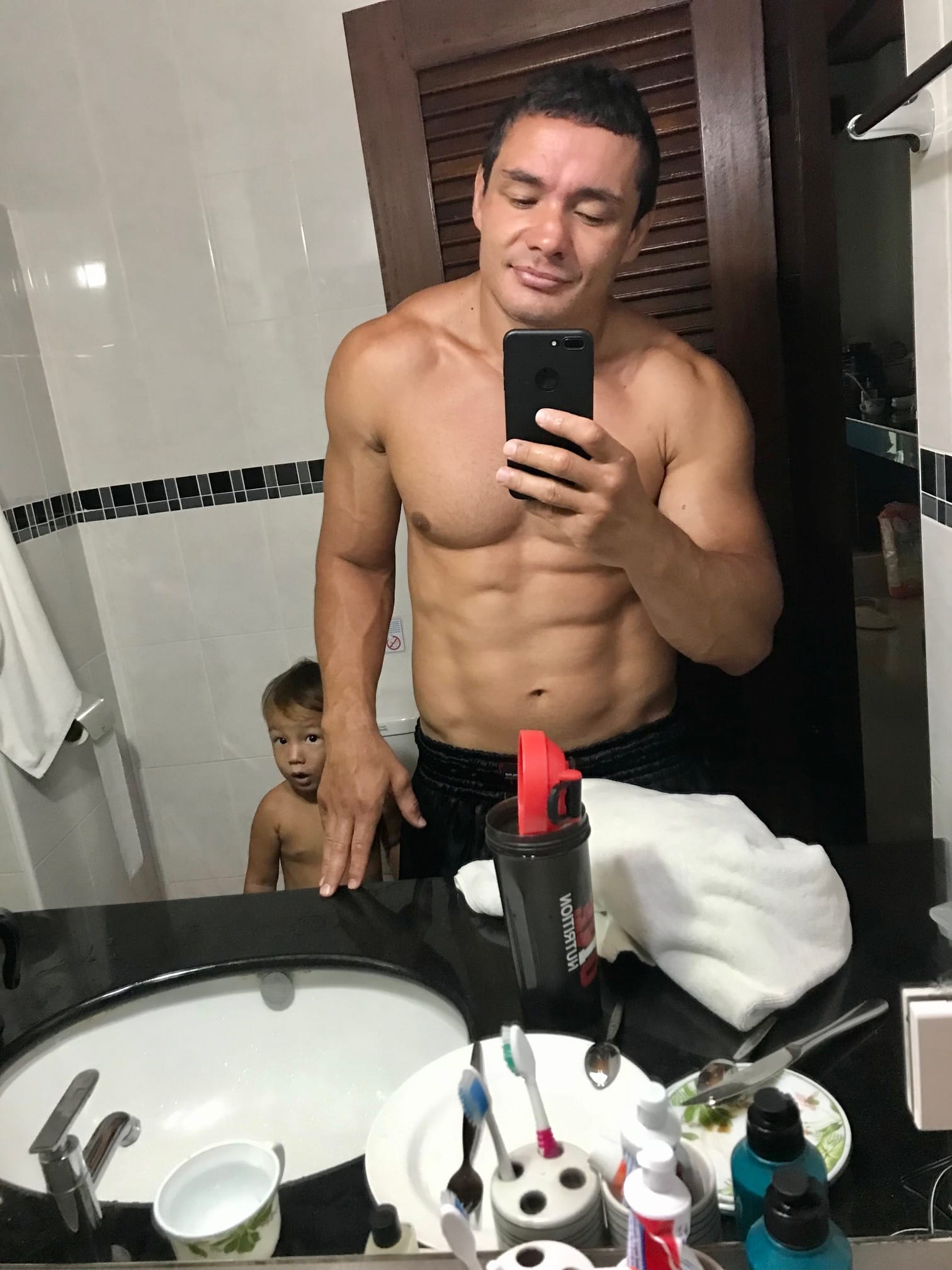 A shirtless man taking a mirror selfie in a bathroom with a small child peeking out from behind him Various toiletries and personal items are on the countertop