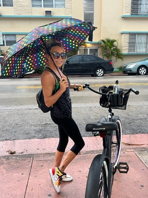 A woman stands beside a bicycle on a sidewalk holding a colorful polka dotted umbrella She is wearing sunglasses a black top black leggings and white sneakers with red accents
