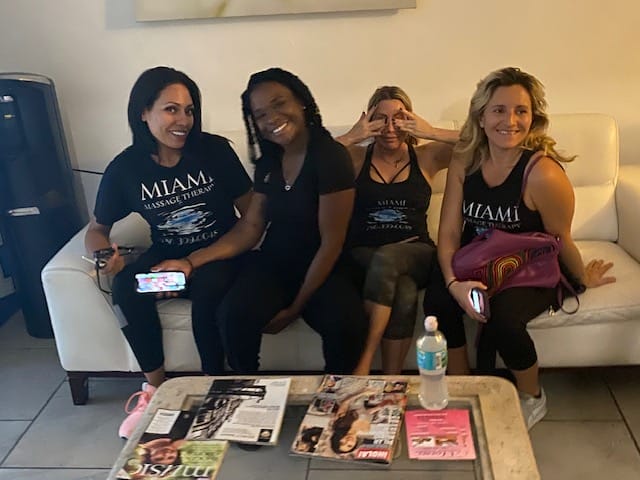 Four women are seated on a white couch with magazines and a water bottle on a table in front of them They are all smiling and appear to be posing for the photo | Miami Massage Therapy Best Female massage therapists