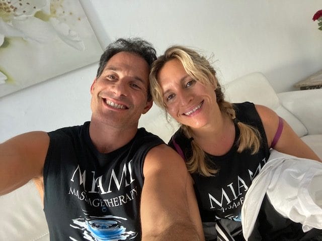 Two individuals wearing matching Miami Massage Therapy shirts smile at the camera seated on a white couch with a flower painting in the background