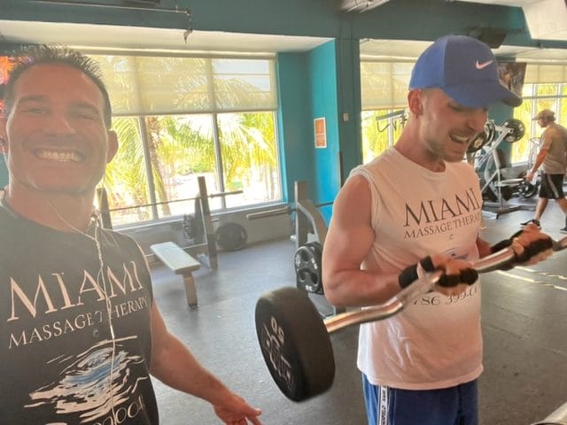 Two men in Miami Massage Therapy shirts are at the gym One is smiling at the camera while the other is lifting weights Exercise equipment and large windows in the background