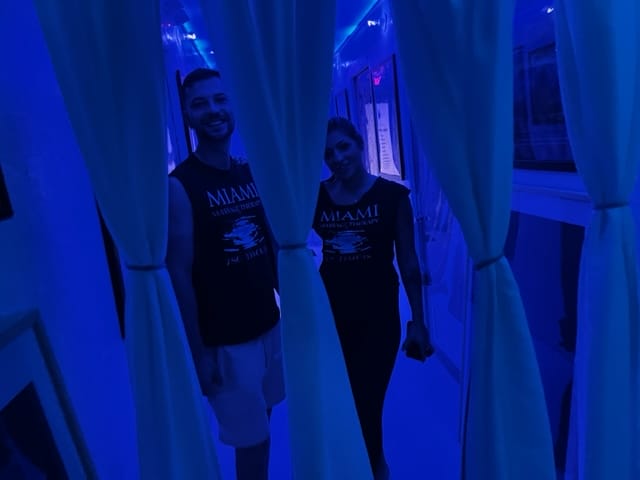 Two people wearing black Miami t shirts stand behind partially drawn white curtains in a blue lit room Both are smiling and looking at the camera